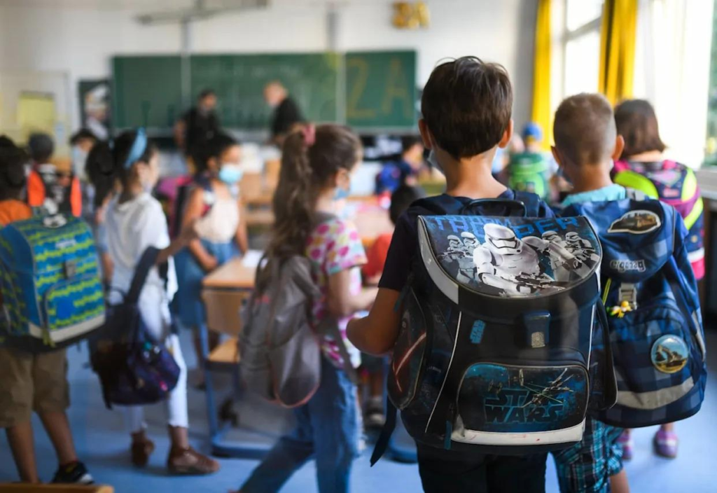 several kids with backpacks in a classroom facing the chalkboard